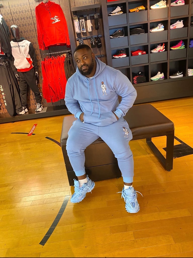Man in light blue tracksuit sitting on brown bench in sneaker store with wall of displayed shoes behind him