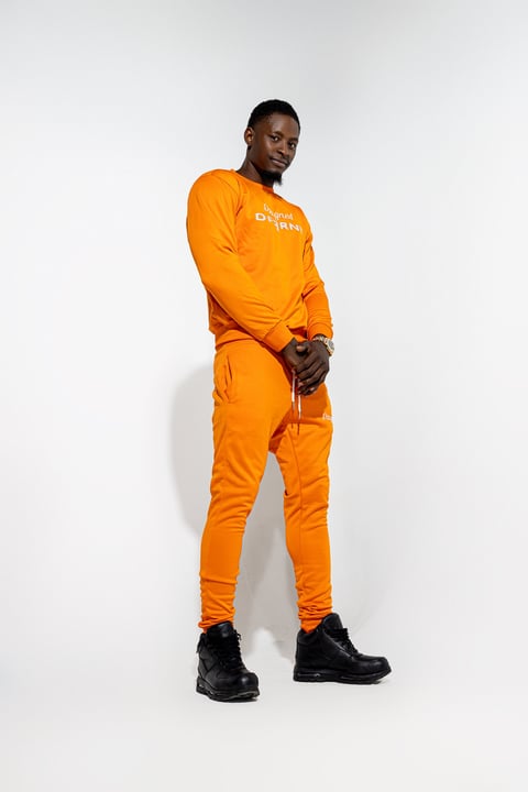 Man in orange athletic tracksuit with white text, black gloves and boots, posing against white background