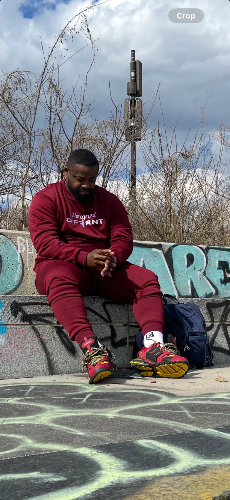 Man in maroon tracksuit sitting on graffiti wall with colorful sneakers, bare trees and cell tower in background