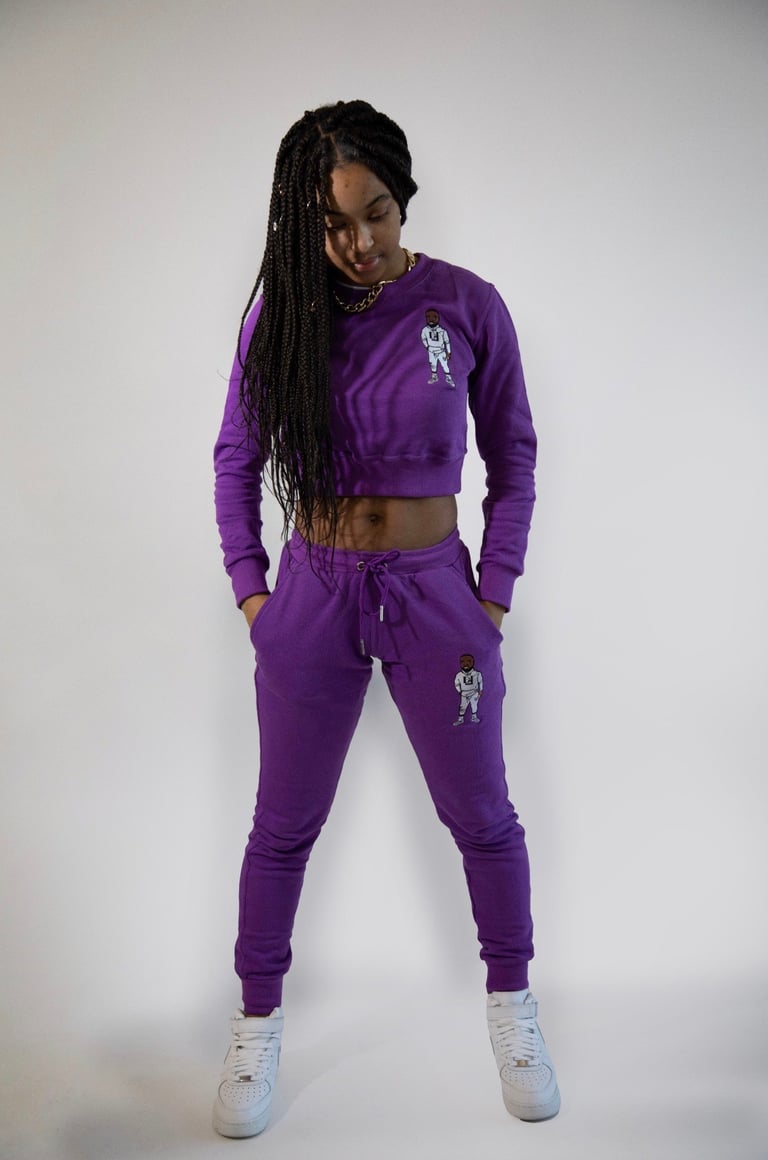 Woman wearing purple cropped sweatshirt and joggers with astronaut embroidery, white sneakers, posing against white background