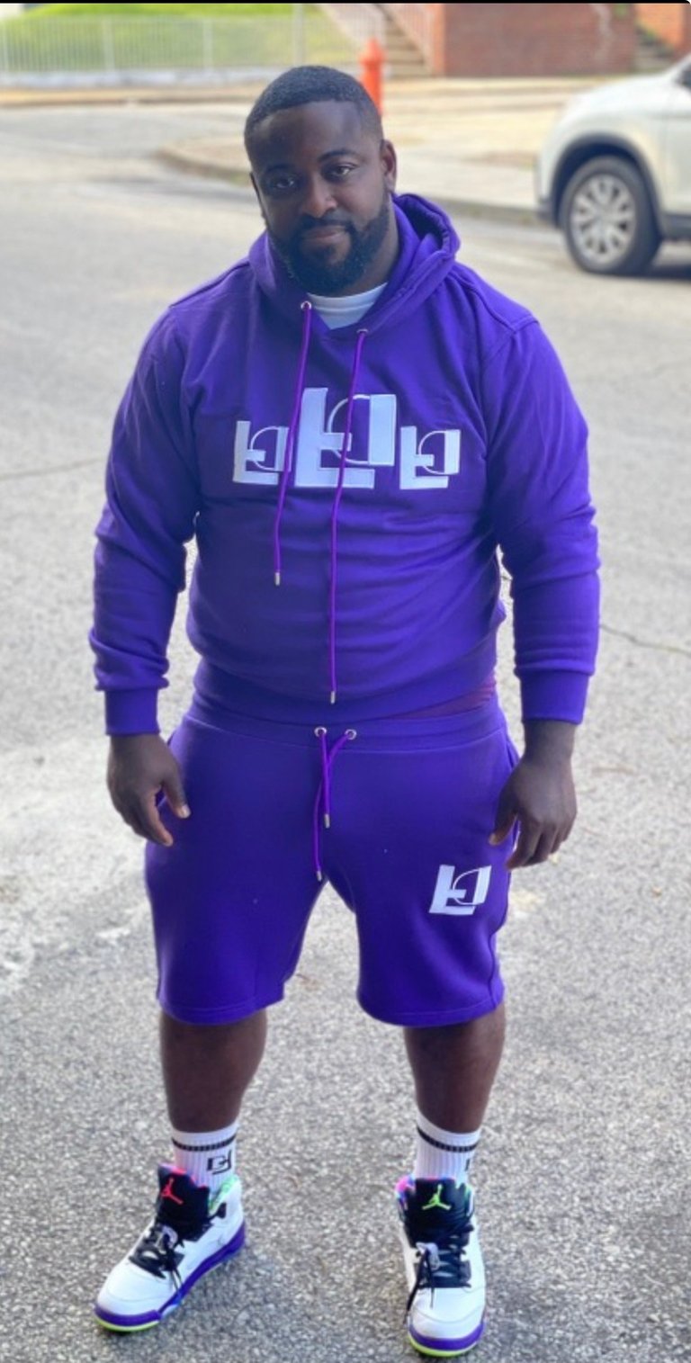 Man wearing purple tracksuit with logo design, white and purple sneakers, standing in parking lot