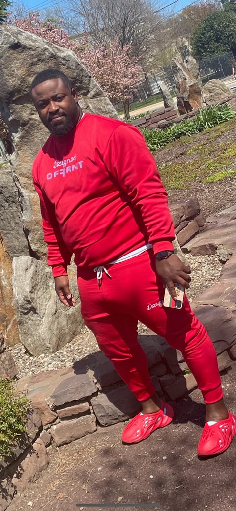 Man wearing a red tracksuit and red sneakers posing on stone steps in a garden with blooming trees and stone walls in the background