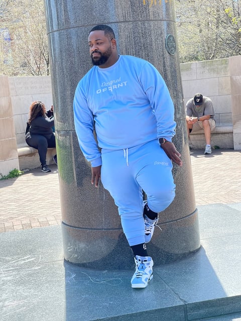 Man in blue tracksuit leaning against a concrete pillar in an outdoor public space with other people in the background